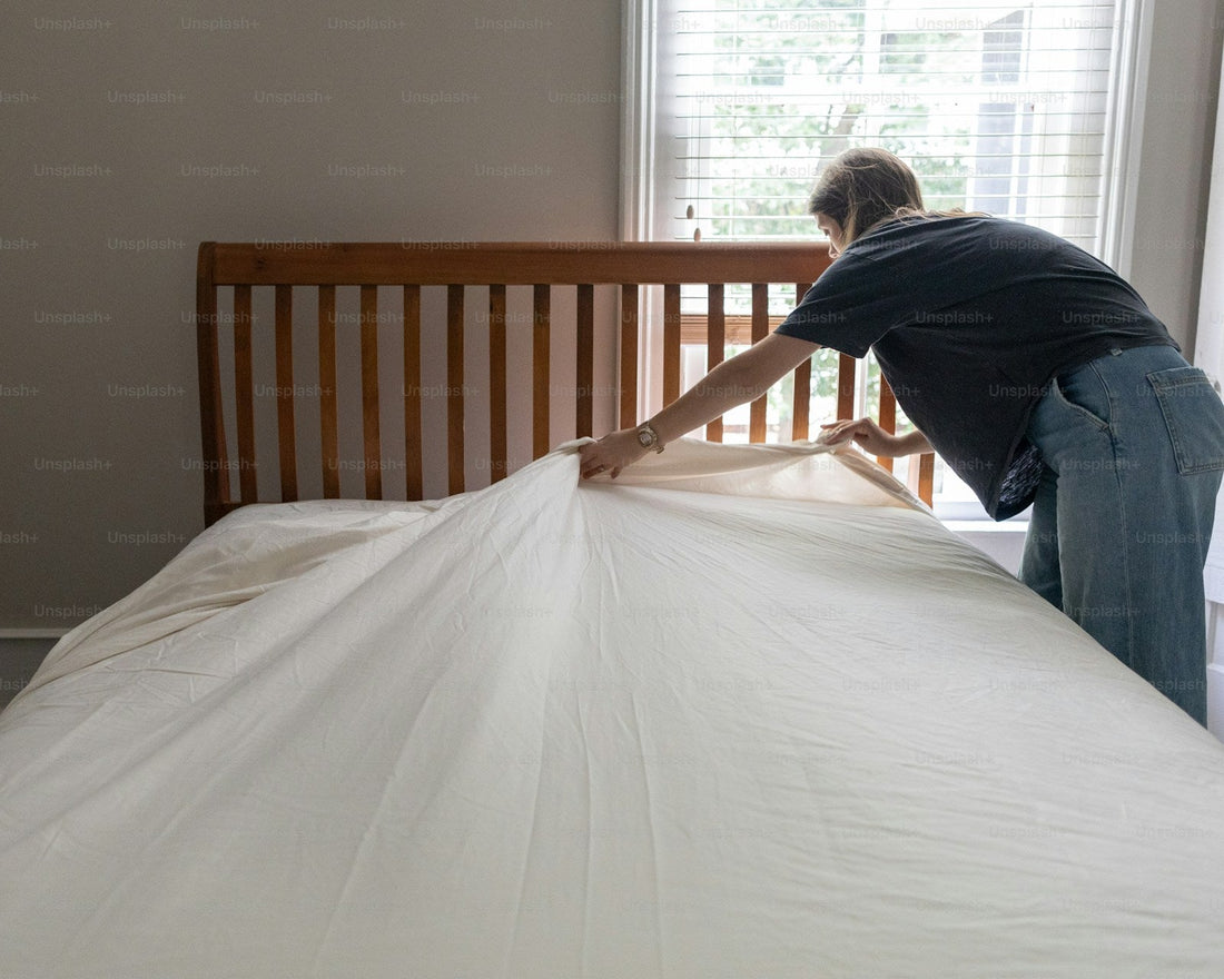 Person placing a clean white sheet over a mattress in a bright bedroom